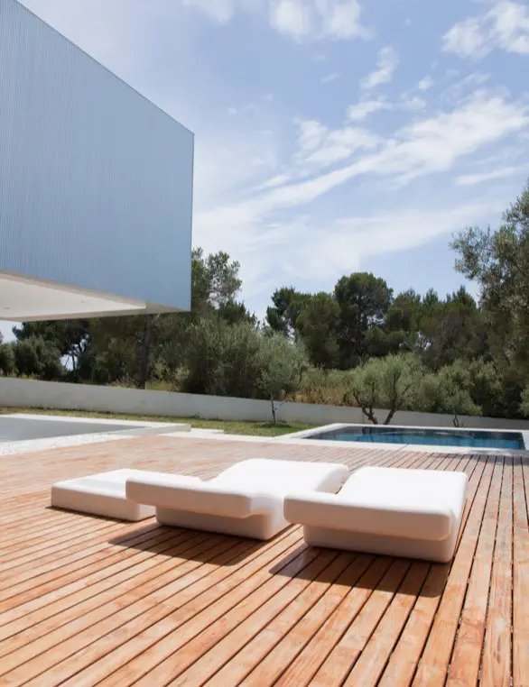White lounge chairs on a wooden deck by a pool with trees in the background.