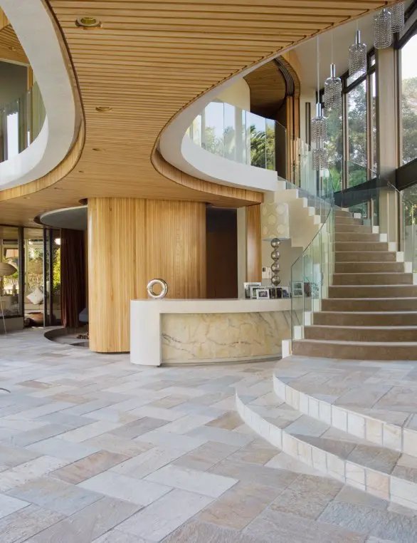 Modern interior of a luxury home featuring a curved wooden ceiling, stone tiled floor, and a grand staircase with a glass